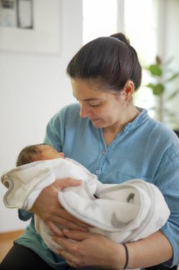 Anne, yeni doğmuş bebeğini yumuşak bir battaniyeye sarıp erken anneliğin huzurlu ve besleyici ortamını ve anne ile çocuk arasındaki derin bağı vurguluyor.
