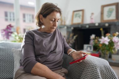 Gri gömlekli, kısa saçlı yaşlı bir kadın desenli bir kanepede oturuyor, ciddi bir ifadeyle kırmızı akıllı telefonuna bakıyor, rahat ve iyi dekore edilmiş oturma odasıyla çevrili.