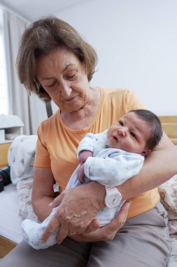 Büyükannem yeni doğan torununu dikkatlice kucağına aldı, şefkatli bir ifadeyle aşağıya baktı, nesiller arasında huzurlu bir ortamda paylaşılan derin bağlantıyı ve şefkati vurguladı.
