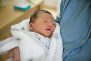 Anne, yeni doğmuş bebeğini yumuşak bir battaniyeye sarıp erken anneliğin huzurlu ve besleyici ortamını ve anne ile çocuk arasındaki derin bağı vurguluyor.