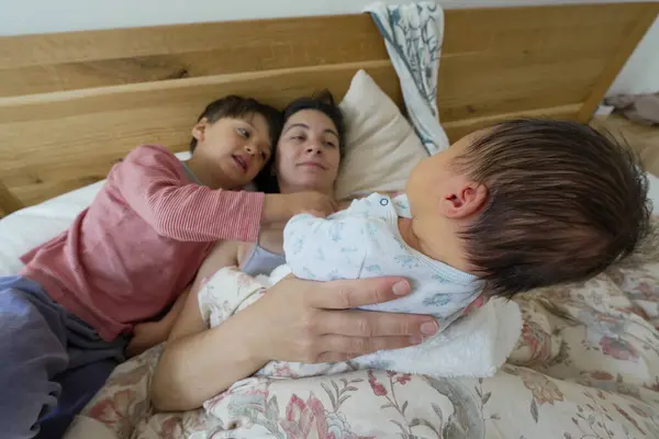 stock image Mother lying in bed with her newborn baby and older child, interacting lovingly, highlighting the family bond and the gentle care shared among them in a warm, home environment