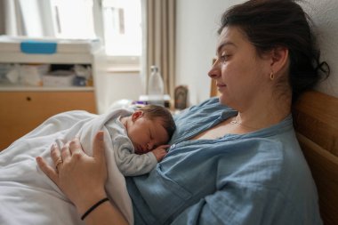 Yeni bir anne sevgiyle yeni doğmuş bebeğini yatağa mavi bir gömlek giyerek yatırır. Bu hassas an anne ve çocuk arasındaki güçlü bağı vurgular nitelikte..