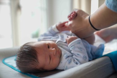 Anne bezini değiştirirken yeni doğmuş bebek soyunma masasında uzanmış merakla bakıyor. Şu anda günlük ebeveynlik görevlerinde gösterilen özen ve özen gösteriliyor