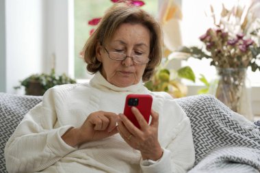 Gözlüklü ve beyaz kazaklı yaşlı kadın bir kanepede oturuyor, akıllı telefonuna odaklanmış. Özenli ifadesi, rahat ve aydınlık bir odada dijital bir etkileşimi vurguluyor.