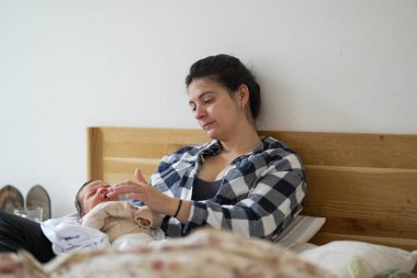 Anne ekose gömleğiyle yatakta oturuyor, bebeğinin minik elleriyle nazikçe oynuyor. Bej bir kıyafet giymiş olan bebek, merakla başını kaldırıp anne-çocuk etkileşiminin ilk anlarını yakalıyor.