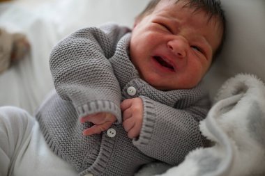 Gri örgü bir kazakla ağlayan yeni doğmuş bir bebek, yumuşak beyaz bir battaniyenin üzerinde yatıyor. Bebeğin koyu renk saçları var ve görünen o ki üzgün, çocukluk döneminin doğal duygularını açık bir şekilde gösteriyor..