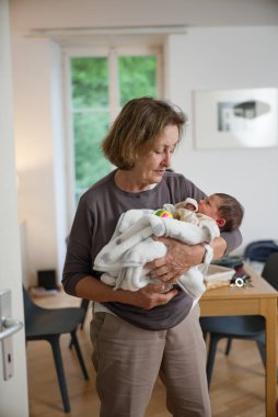 Büyük anne beyaz bir battaniyeye sarılı yeni doğmuş bir bebeği kucağında tutuyor, bebeğe şefkatle bakıyor. Konforlu ev dekorasyonu, sahnenin sıcak ve besleyici atmosferini arttırır..