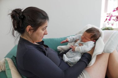 Anne sevecen bir şekilde yeni doğmuş bebeğini bir koltukta otururken kucaklıyor, şefkatle ve dikkatle bebeğe bakıyor, yumuşak ışıklandırmalı ve rahat bir evde oturuyor.