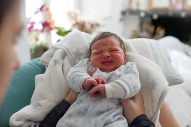 Yeni doğan bebek, kucağında mavi desenli bir tulum, sıcak bir ev ortamında yoğun bir şekilde ağlıyor. Bebeğin ifadesel yüzüne ve ellerine odaklanıyor ve arka planı bulanık.