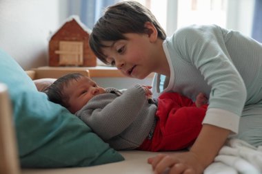 Büyük kardeş yeni doğan bebeğin üzerine eğiliyor, bebekle nazikçe bağ kuruyor. Pencereden gelen parlak doğal ışık sevgi dolu bağı ve sıcak aile anını güçlendirir.