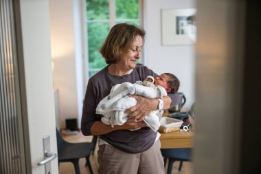 Büyük anne beyaz bir battaniyeye sarılı bebeği kucağında tutuyor, gülümseyerek bebeğe bakıyor. Ortam sıcak ve davetkar bir ev ortamı, jenerasyonun dokunaklı bir anını yakalıyor.