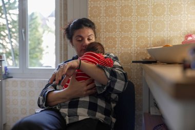 Anne, sarı desenli duvar kağıdıyla sıcak bir mutfakta oturan kırmızı ve beyaz çizgili elbiseli bir bebeği kucağında tutuyor. Annenin nazik kucaklaması ve bebeklerin rahat duruşu.