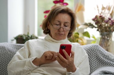 Gözlüklü ve beyaz kazaklı yaşlı kadın bir kanepede oturuyor, akıllı telefonuna odaklanmış. Özenli ifadesi, rahat ve aydınlık bir odada dijital bir etkileşimi vurguluyor.