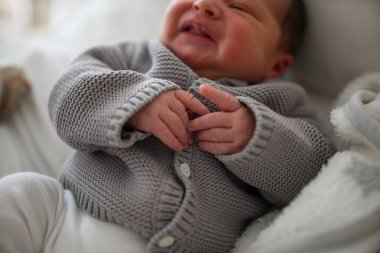 Gri bir hırka ve beyaz pantolon içinde yeni doğmuş bir bebek, yumuşak bir yatakta uzanıyor, hafifçe gülümsüyor ve halinden memnun görünüyor, rahat bir ev ortamında, yumuşak ışıklandırma ve rahatlatıcı bir dekorla