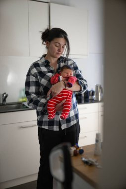 Yeni doğmuş bebeğini kırmızı beyaz çizgili bir elbiseyle bir mutfakta tutan bir annenin yakın çekimi. Bebeğin sakin ifadesi ve annenin sevgi dolu bakışları huzur verici ve iç açıcı bir bakış oluşturur.