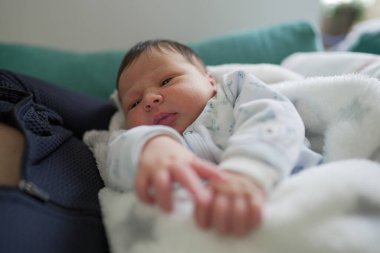 Yumuşak beyaz bir battaniyeye sarılı yeni doğmuş bir bebek dikkatlice etrafına bakıyor. Bebek şirin, desenli bir tulum giymiş hassas ve meraklı bir anı yakalıyor.
