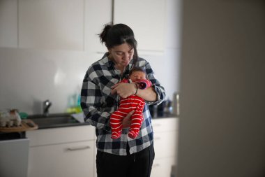 Mother in checkered shirt holding newborn in a red and white striped outfit, standing in a bright kitchen. The baby is cradled in her arms, eyes half-open, creating a peaceful and tender family moment clipart