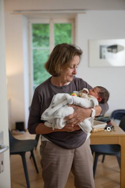 Büyükannesi yeni doğan torununu yumuşak beyaz bir battaniyeye sarmıştı. Bebeğe sevgiyle bakıyor, sıcak ve samimi bir aile ortamı yaratıyor..