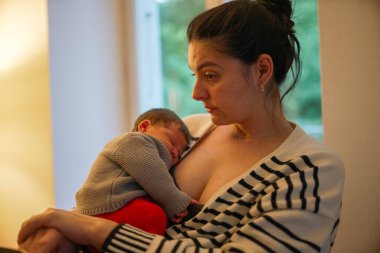 Bej kıyafetli yeni doğmuş bir bebek anne babanın göğsünde dinleniyor ve tuhaf bir ifadeyle etrafa bakıyor. Samimi an, ebeveyn ve çocuk arasındaki bağı vurgular, yumuşak ışıklandırma sahneyi güçlendirir.