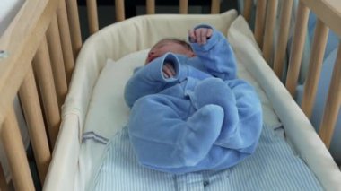 Mavi kıyafetli yeni doğmuş bir bebek hastane odasındaki tahta bir karyolada huzur içinde uyuyor. Tıbbi ortamda yeni doğanlara sağlanan huzur ve bakımı vurguluyor.