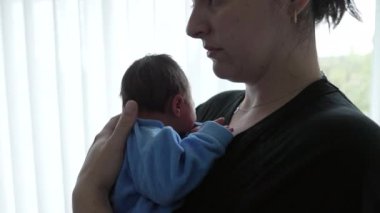 Mother holding newborn baby in a blue onesie, side profile, close embrace, showing love and care, soft background lighting, intimate bonding moment, capturing early days of motherhood and infancy