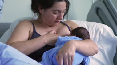 Anne yeni doğmuş bebeği mavi tulumla emziriyor, hastane yatağında oturuyor, doğum sonrası bakım ortamında anne ve çocuk arasında özel ve şefkatli bir an sergiliyor.