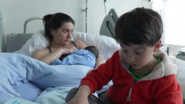 Mother breastfeeding newborn while older sibling in red jacket sits nearby on hospital bed. Mother's expression shows fatigue, and child plays by himself. Family bond in hospital