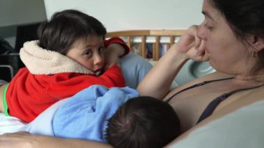 Mother breastfeeding newborn while older child leans on her, conveying the warmth and closeness of family life and the support that older siblings provide during early parenting