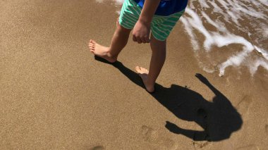 Çocuk sahilde yürüyor, okyanusa bakıyor, dalgalar kıyıya vuruyor.