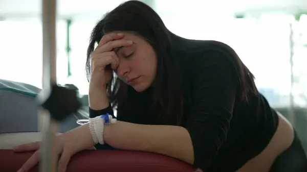 stock image Pregnant woman suffering from contractions during pre labor at hospital clinic covering face as she leans on bedside, IV drip attached in hand, real life struggling expression