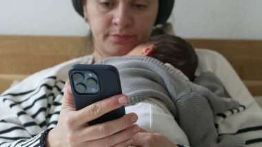 Annenin kucağında yeni doğmuş bir bebeğin göğsünde dinlenirken cep telefonu cihazını tutarken, bebeğin hayatının ilk haftasında yatağa uzanırken yakın çekimi. Telefon ile çevrimiçi içeriği tarayan kişi