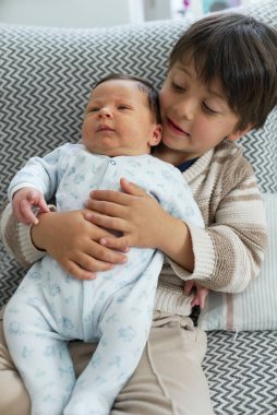 Büyük kardeş ve yeni doğan kardeş desenli bir yastıkta oturup aile sevgisinin tatlı bir anını yakalıyorlar. Bebek mavi tulumlu, ve kardeşi çizgili süveter giyiyor.
