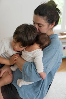 Anne yeni doğmuş bebeğini sakinleştirirken büyük çocuğu ona bakıyor, hassas bir aile anını yakalıyor. Sahne, aile içindeki sevgi ve beslenme bağının sakin bir ortamda vurgulandığını gösteriyor.