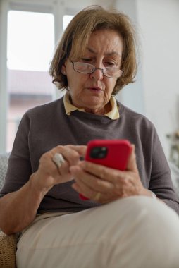 Yaşlı kadın akıllı telefonuna odaklandı, modern teknolojiyi kullanarak, doğal ışıkla iç mekan sahnesi, yaşlı vatandaşlar arasındaki dijital bağlılık ve teknolojik açıklığı vurgulayarak, rahat ev ortamları
