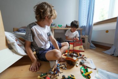 Samimi bir odada yerde oyuncaklarla oynayan iki çocuk. Büyük oğlan küçük olana bakıyor, neşeli etkileşimlerini ve kardeş bağlarını parlak, neşeli bir ortamda vurguluyor..