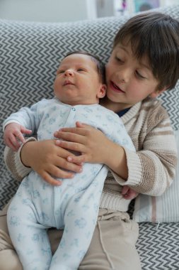 Ağabeyi yeni doğan kardeşini nazikçe tutuyordu. İkisi de desenli yastıktaydı. Bebek mavi bir tulum giyerken, erkek kardeş çizgili bir kazak giyiyor.,
