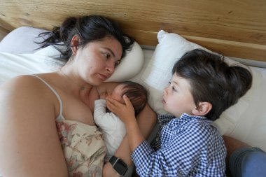 Anne yeni doğmuş bebeği emzirirken büyük çocuk sevgiyle bebeğin başını tutar, aile birliği, rahat ortam, annelik sevgisini yakalar ve kardeşlik bağı kurar, huzurlu bir ortam.