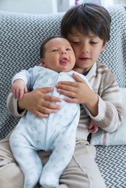 Neşeli ağabey, yeni doğan kardeşini rahat bir koltuğa oturtup, samimi bir aile anını yakalıyor..