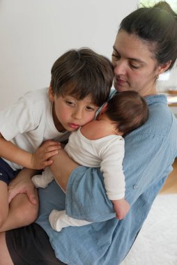 Anne büyük çocuğunu teselli ederken yeni doğmuş bir bebeği kucağında tutuyor. Bu samimi aile anı huzurlu bir ev ortamında anne sevgisi, bağlılık ve şefkat gösteriyor..