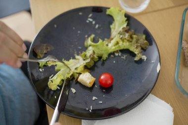 Salata artıkları ve kiraz domatesli siyah tabağın yakın çekimi, yemeğin sonunu gösteriyor, basit, sağlıklı yeme alışkanlıklarını ve günlük yemek düzenini temsil ediyor.