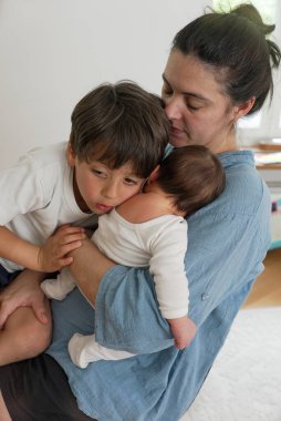 Anne yeni doğmuş bebeğini kucaklıyor ve büyük çocuğunu samimi bir aile ortamında teselli ediyor. Bu sahne anne sevgisinin özünü yansıtıyor ve huzurlu bir ev ortamında bağ kuruyor..