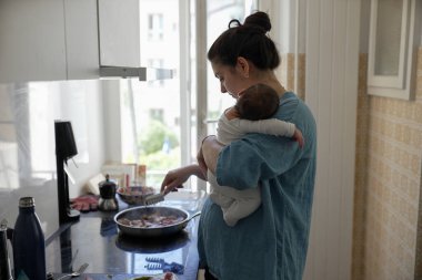 Anne mutfakta yemek pişirirken bebeğini kucağında tutuyor, çocuk bakımı ve yemek hazırlama işini dengelemek için çoklu görev yapıyor, modern bir annenin gündelik yaşamını resmediyor aile ve ev işlerini yönetiyor.