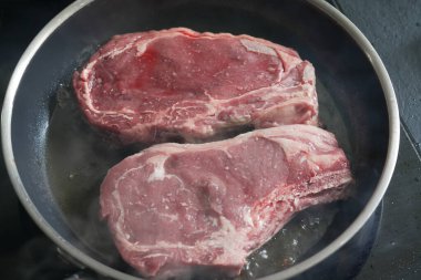 Sıcak tavada kızaran iki çiğ pirzola, buhar yayıyor, pişirmenin ilk aşamalarını yakalıyor, yemek hazırlamaya elverişli, ve biftek tarifi içeriği...