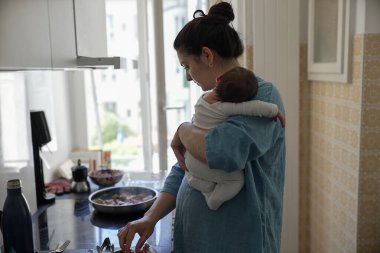 Anne, modern mutfakta yemek pişirirken bebeği kucağına alır, yemek hazırlar ve ebeveynlik görevlerini dengeler, ev sorumluluklarını yöneten çok görevli bir annenin günlük yaşamını temsil eder.