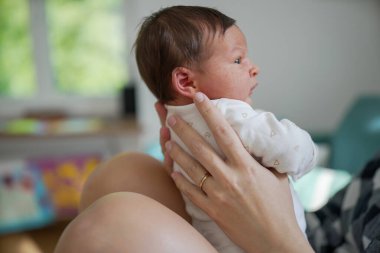 Anne kucağında yeni doğmuş bir bebeğin yan görüntüsü, ikisi de birbirine bakıyor, sevgi dolu ve sakin bir atmosferle, anne ve bebek arasındaki özel bağ ve bağın altını çiziyor.