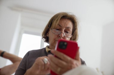 Akıllı telefon kullanan yaşlı kadın, ekrana odaklanıyor, doğal ışıkla iç mekan sahnesi, yaşlılar arasında dijital bağlılık ve modern teknoloji kullanımını yakalıyor, rahat ev atmosferi