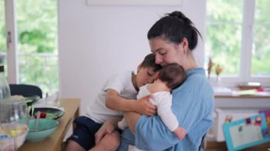 Anne hem bebeği hem de küçük bir çocuğu kucağında tutuyor, sıcak bir şekilde gülümsüyor. Kapalı bir ortamda ebeveynliğin neşeli ve besleyici tarafı.