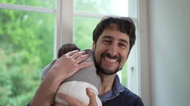Yeni doğmuş bebek babasının omzunda dinleniyor, rahat örülü bir kazak giyiyor. Baba pencereye doğru döner, aralarındaki rahatlık ve güven dolu kucaklaşmayı vurgular. 
