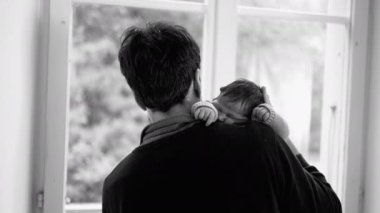 Baba yeni doğmuş bebeği nazikçe omzundan tutup pencereden dışarı bakıyor, aile sevgisi, siyah beyaz, sakin bir sahne, huzurlu bir sabah, ebeveyn-çocuk bağı.