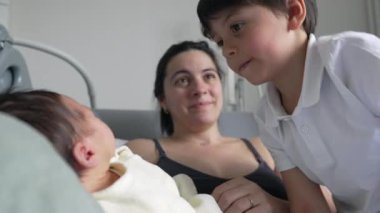 Küçük çocuk yeni doğmuş bir bebekle nazikçe etkileşime giriyor, alınları sevgi dolu bir jestle birbirine dokunuyor. Anne ve küçük oğlu hastanede ailenin yeni üyesiyle kaynaşıyor.